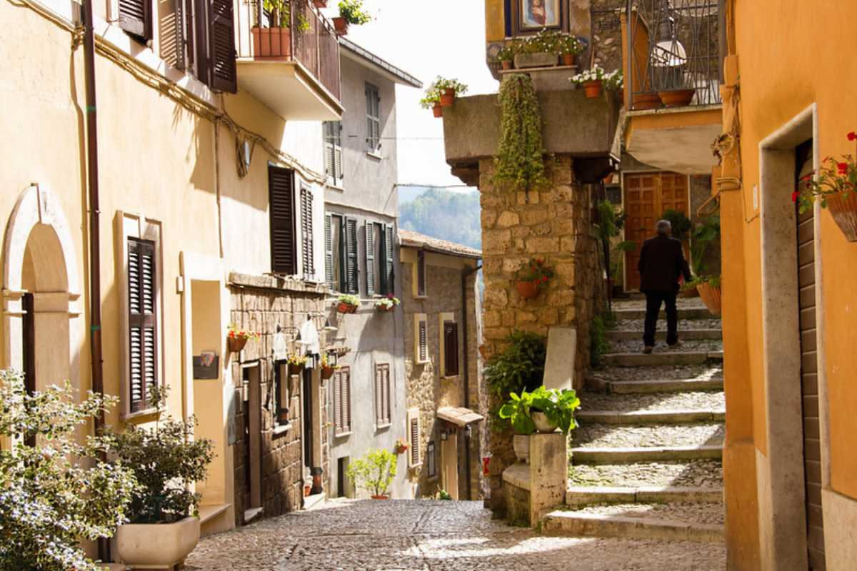 Piazza centrale di un borgo italiano con case in pietra, strada in selciato, campanile sullo sfondo e abitanti che camminano tra vicoletti stretti
