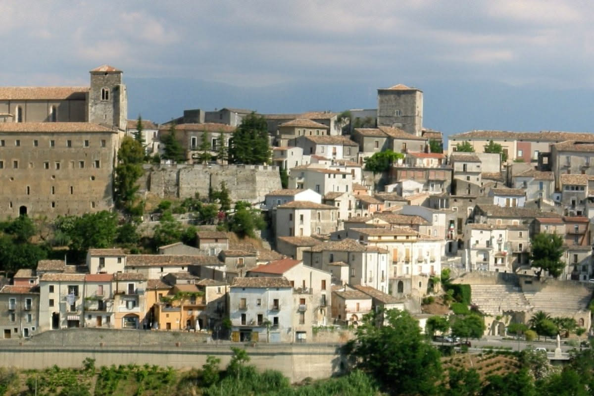 Strada acciottolata di un borgo italiano montano con case in pietra, una piazza con una fontana e anziani che parlano seduti su panche di legno