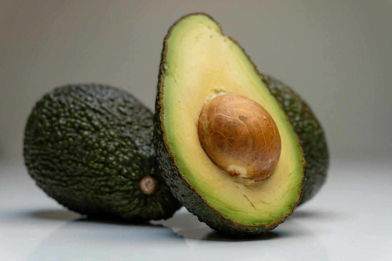 Mezzo avocado tagliato fresco con il nocciolo visibile, posto su un tagliere di legno accanto a un coltello d'acciaio, con foglie verdi di sfondo