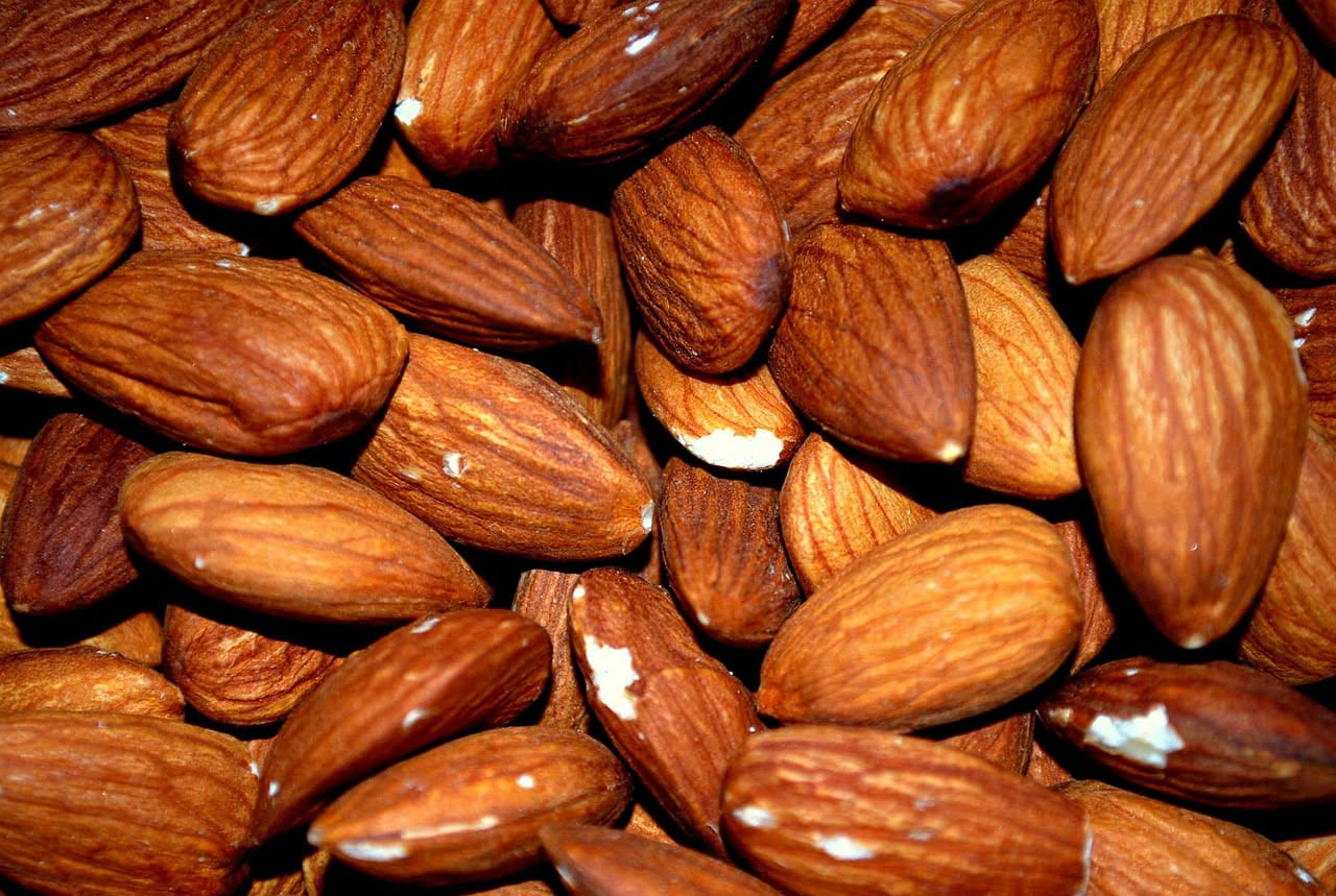Mandorle sgusciate e intere in una ciotola di legno chiaro, su sfondo bianco, con alcune mandorle sparse accanto