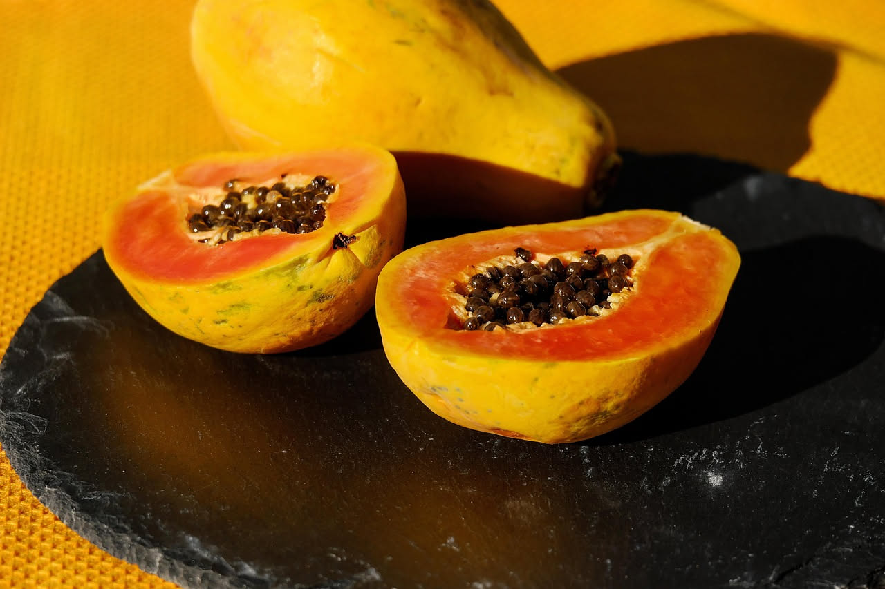 Papaya gialla intera e tagliata in metà con semi neri visibili, su sfondo di foglie verdi e tavolo di legno naturale