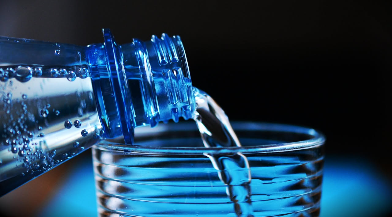 Bicchiere d'acqua trasparente con cubetti di ghiaccio e limone su sfondo bianco, con gocce di condensa e sole brillante sullo sfondo sfocato