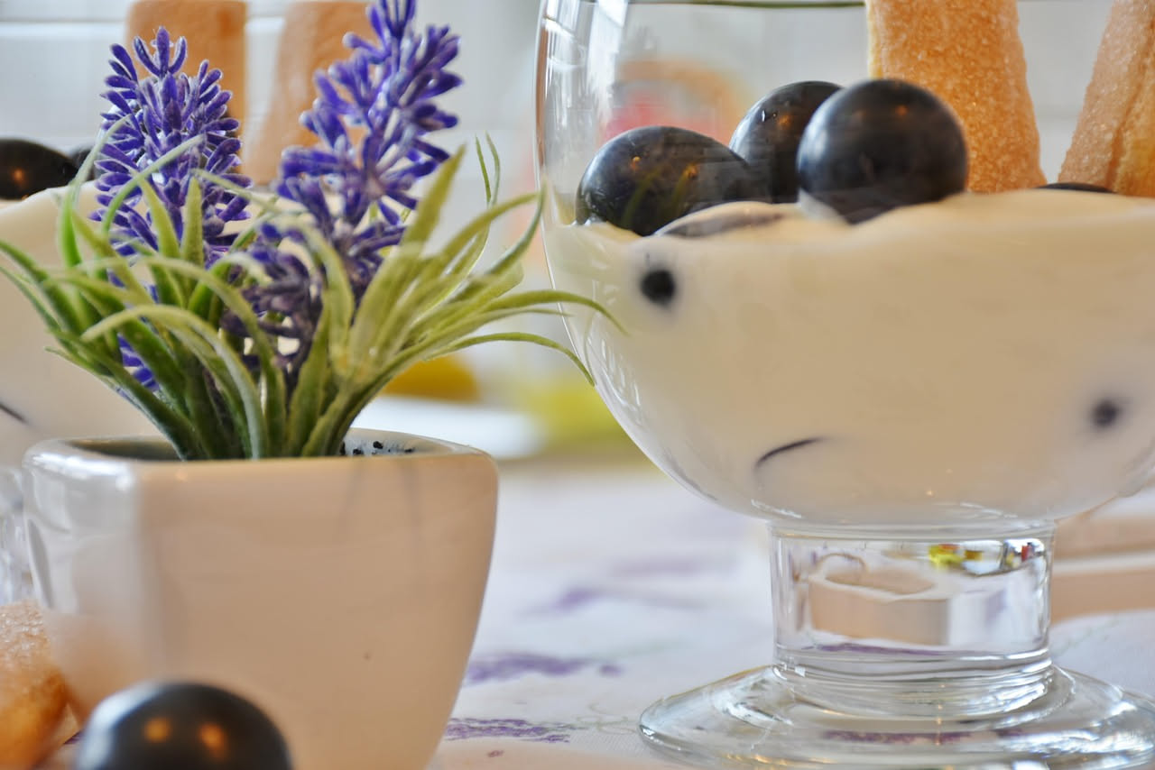 Ciotola di yogurt greco bianco con frutta fresca estiva, miele e granola su un tavolo di legno al sole, con bicchiere d'acqua accanto