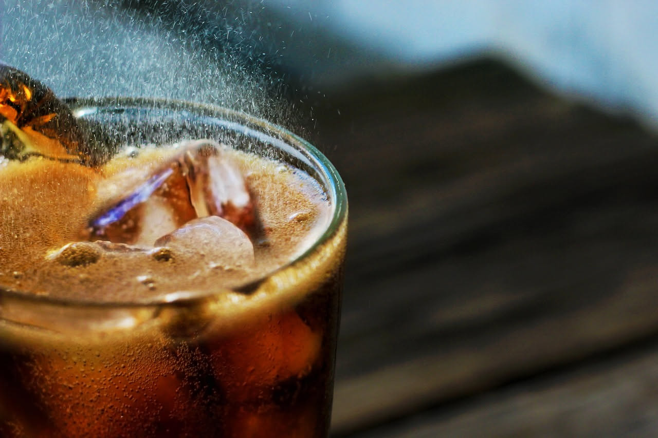 Bicchiere alto con caffè freddo marrone, cubetti di ghiaccio, schiuma di latte e cucchiaino metallico su tavolo bianco con sfondo naturale sfocato