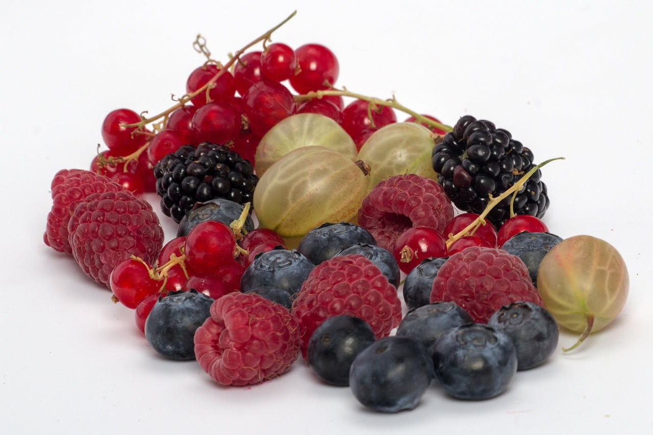 Ciotola di ceramica bianca colma di fragole rosse, mirtilli blu, lamponi rossi e more nere su un tavolo di legno, con una finestra luminosa sullo sfondo che illumina i frutti di bosco
