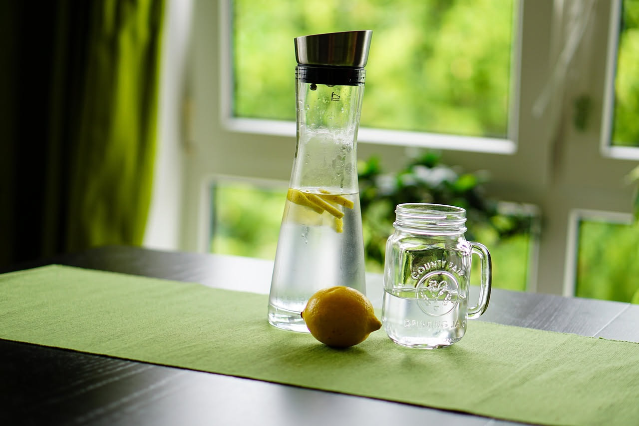Bicchiere di vetro trasparente con acqua fresca e fetta di limone giallo su sfondo bianco, raggi di sole che illuminano la bevanda